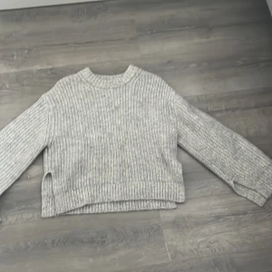 Grå ribbstickad tröja - Säljer en grå ribbstickad tröja med rund hals och långa ärmar. Tröjan har en loose passform och är perfekt för kyliga dagar. Den är enkel att matcha och har en stilren look med diskreta slitsar i sidorna.