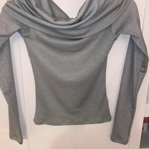 Glittrig silver topp från H&M Divided - Snygg långärmad topp i glittrigt silver från H&M Divided. Toppen har en bred, offshoulder med urringad hals. Perfekt för dig som vill sticka ut med lite extra shine. Materialet är stretchigt och sitter snyggt på kroppen. Aldrig använd.