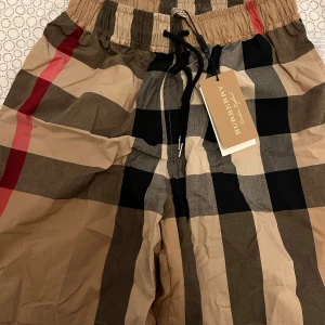 Burberry rutig shorts med snörning - Snygga Burberry shorts i klassiskt rutigt mönster med beige, svart, brun och röda toner. Modellen har elastisk midja med svart snörning och är tillverkad i 100% bomull för skön känsla. Perfekt för dig som vill ha en ikonisk streetstyle-look.