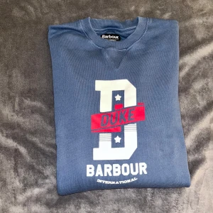 Blå sweatshirt från Barbour International - Snygg blå sweatshirt från Barbour International med stort tryck framtill där det står 'DUKE' och 'BARBOUR'. Tröjan har rund hals, ribbade muddar och är långärmad. Materialet känns som mjuk bomull och polyester, perfekt för chill dagar.