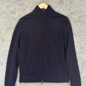 Blå merinoullskofta från These Glory Days - Blå kofta i merinoull från These Glory Days. Stilren design med hel dragkedja framtill, hög krage och ribbade muddar. Perfekt för lager-på-lager och funkar året runt. Snygg och enkel att matcha med allt.