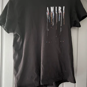Svart AMIRI t-shirt med färgstänk - Svart t-shirt från AMIRI i 100% ekologisk bomull. Framsidan och baksidan har AMIRI-logga med coola färgstänk i blått, vitt, rött och orange som rinner ner över tyget. Klassisk passform och korta ärmar. Skriv vid minsta fråga priset är inte hugget i sten!💯