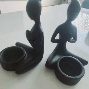 Statyer  - Bilderna visar två dekorativa figurer i svart, sittande i meditationsställning med skålar framför sig. De är stilrena och passar som ljushållare eller dekoration. Inga synliga defekter eller större slitage. Perfekt för att skapa en lugn atmosfär i hemmet.