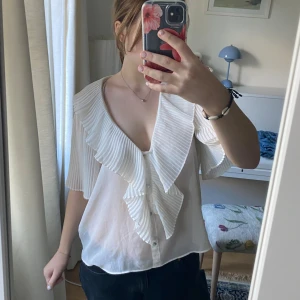 Vit blus med volang och knappar - Säljer en genomskinlig luftig vit blus med djup v-ringning, stora plisserade volanger längs kragen och framtill. Knappar framtill och korta ärmar. Tunt, transparent tyg som känns lätt och somrigt. Lite tomatfläclar och ett litet hål men syns inte på avstånd💕