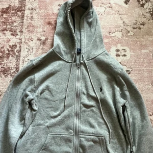 Grå hoodie från Polo Ralph Lauren - Grå hoodie från Polo Ralph Lauren med dragkedja framtill, huva med snörning och klassisk broderad logga på bröstet. Tröjan har känguruficka och är tillverkad i mjuk bomullsblandning. Perfekt för chill dagar eller när du vill ha en clean look.