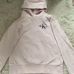 Ljusrosa hoodie från Calvin Klein - Säljer en ljusrosa hoodie från Calvin Klein Jeans med broderad logga i blått och svart på bröstet. Tröjan har huva, långa ärmar och ribbade muddar. Perfekt för en chill och stilren look.