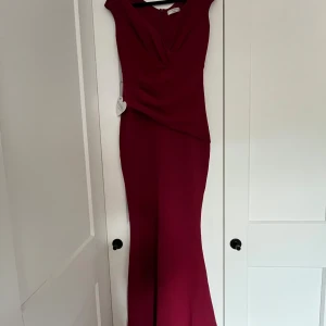 Vinröd långklänning med drapering - Supersnygg vinröd långklänning från Chi Chi London med draperad överdel och figurnära passform. Klänningen har breda axelband och en elegant, lätt utsvängd nederdel. Perfekt för bal eller fest. Materialet känns mjukt och följsamt.
