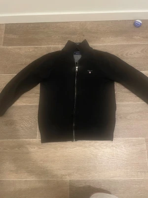 Svart stickad kofta från GANT - Svart stickad kofta från GANT med hel dragkedja framtill och broderad logga på bröstet. Klassisk och stilren modell med ribbade muddar och hög krage. Perfekt för lager-på-lager och passar till det mesta.