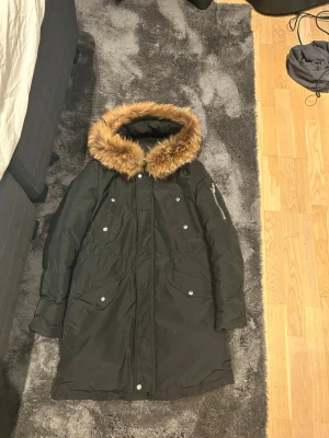 Svart Rockandblue parkas med päls - Svart parkas från Rockandblue med snygg pälsimitation runt huvan. Jackan har quiltat foder, dragkedja och flera fickor med knappar. Perfekt för kalla dagar och har en stilren look med detaljer på ärmen.