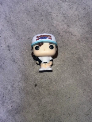 Ingen väst synlig på bilden - Dustin from stranger things. Bilden visar en liten figur med keps och vit skjorta, men ingen väst är synlig eller kan beskrivas utifrån bilden.