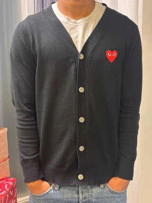 Svart kofta Comme des Garçons Play S - Svart kofta från Comme des Garçons Play i storlek S. Klassisk design med v-ringning, knappar framtill och ett rött hjärta med ögon broderat på bröstet. Mjuk och skön kvalitet, perfekt att slänga på över en t-shirt. Snyggt ribbade muddar vid ärmslut och nederkant.