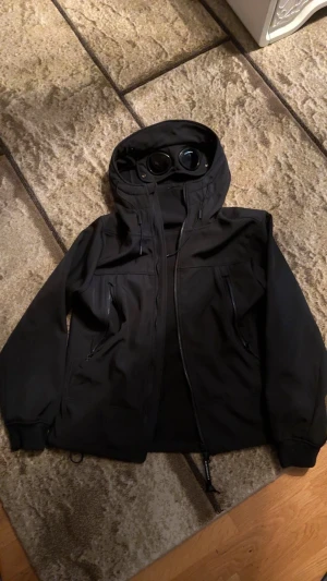 C.P. Company jacka  - Säljer en svart Goggle Jacket från C.P. Company med ikoniska glasögon i huvan. Jackan har två sidofickor med dragkedja, justerbar huva med snörning och är tillverkad i ett slitstarkt syntetmaterial. Perfekt för dig som vill sticka ut med en unik och urban look.