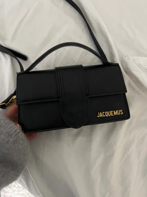 Svart Jacquemus axelväska i skinn - Snygg svart axelväska från Jacquemus med stilren rektangulär form och guldfärgade detaljer. Väskan har ett lock med magnetknäppning och justerbar axelrem. Tillverkad i slätt skinn med Jacquemus-logga i guld på framsidan. Perfekt för dig som gillar minimalistisk design.
