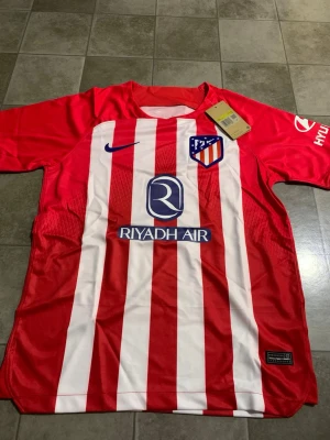 Atletico Madrid randig fotbollströja Nike - Säljer en officiell Atletico Madrid fotbollströja från Nike i storlek S. Tröjan är röd och vit med vertikala ränder, klubbmärke på bröstet och sponsorer som Riyadh Air och Hyundai. Tillverkad i lätt polyester med korta ärmar och rund hals.