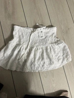 Vit volangkjol med broderade detaljer - Superfin vit kortkjol från H&M med broderade blommor och volangkant. Kjolen har omlottkänsla och knytband i midjan för extra snygg passform. Perfekt för varma dagar och ger en fräsch och somrig vibe.