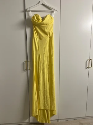 Gul axelbandslös långklänning - Snygg gul långklänning med axelbandslös design och draperad överdel. Klänningen har en elegant och smickrande siluett som faller mjukt längs kroppen. Perfekt för dig som vill sticka ut med en färgstark look. Materialet känns lätt och bekvämt mot huden.