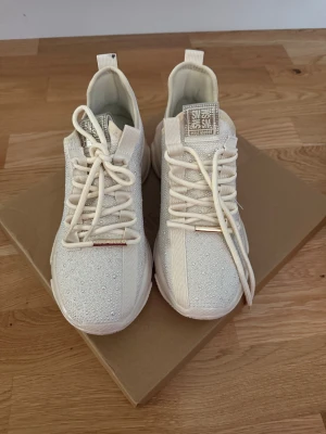 Steve Madden Mysteria-P sneakers pärlvit - Chunky sneakers från Steve Madden i pärlvit färg med glittriga stenar över hela ovandelen. Snygg logga på sidan och plös, snörning framtill och tjock, mönstrad sula. Tillverkade i syntetmaterial och tyg för en modern look.