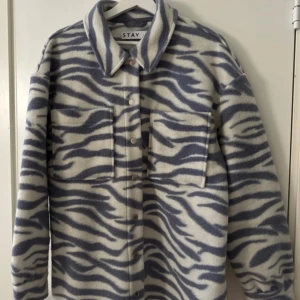 Zebramönstrad overshirt från Stay - Overshirt i mjukt material från Stay med grått och vitt zebramönster. Skjortan har stora bröstfickor, knappar framtill och klassisk krage. Perfekt oversized passform för en trendig look.