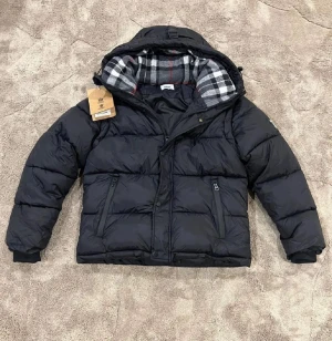 Svart pufferjacka från Burberry - (BYTE BARA)Svart pufferjacka från Burberry med huva och rutigt foder i grått, vitt och rött. Jackan har två dragkedjefickor fram, ribbade muddar och tydlig Burberry London England-patch på ärmen. Perfekt för kalla dagar med sin tjocka och fluffiga design.