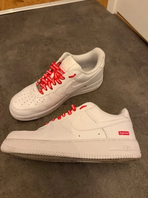 Nike Air Force 1 x Supreme sneakers - Nike Air Force 1 Supreme sneakers i helvitt skinn med röda Supreme-loggade skosnören och Supreme box logo på sidan. Klassisk rund tå, perforerad tåbox och platt sula. Snygg och clean design med streetwear-vibe, perfekt för dig som gillar limited edition och hype.