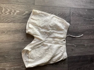 Vita linneshorts med snörning från H&M - Säljer ett par vita shorts från H&M i storlek M. De har en relaxed passform, snörning i midjan och knappstängning. Materialet är mjuk bomull, perfekt för varma dagar. Klassisk design med enkel look som funkar till många stilar.