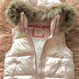 Ljusrosa dunväst med fuskpäls från H&M - Snygg ljusrosa dunväst från H&M med avtagbar huva och fuskpäls. Västen har quiltad design, dragkedja framtill och praktiska fickor. Insidan är fodrad i vitt och rosa, och huvan har fluffig fuskpäls för extra stilpoäng. Perfekt för lager-på-lager under kyliga dagar.