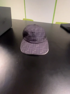 Fendi keps  - Bilderna visar en mörkgrå Fendi-keps med FF-logga, inte en väst. Kepsen har ett rutigt mönster och är tillverkad i ett tygmaterial.