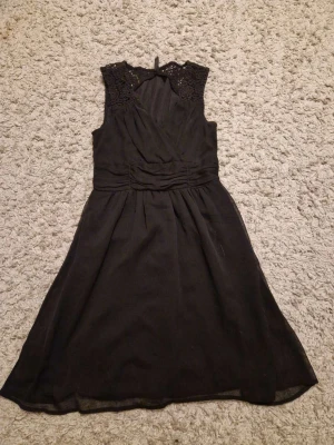 Svart spetsklänning från Vero Moda - Supersnygg svart klänning från Vero Moda med spetsdetaljer vid axlarna och omlottdesign upptill. Klänningen är ärmlös, har markerad midja och lätt utställd kjol. Perfekt för dig som vill ha en klassisk men ändå trendig look.