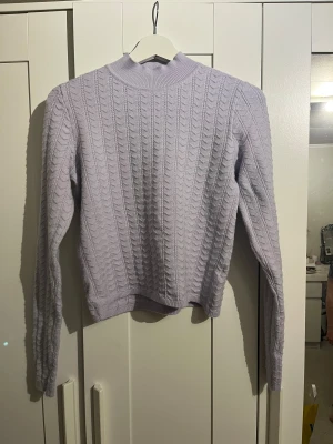 Ljuslila stickad tröja från Lindex - Supersöt ljuslila stickad tröja från Lindex i storlek S. Tröjan har ett snyggt kabelstickat mönster och ribbad hög hals. Perfekt för dig som gillar mjuka och mysiga plagg med långärmad passform och lite kortare längd.