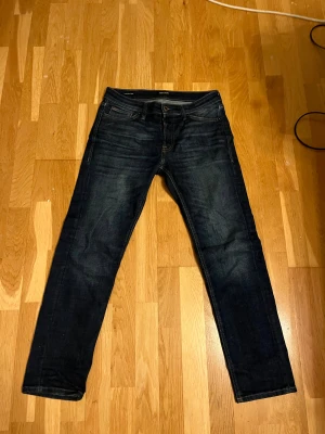 Mörkblå jeans från Jack & Jones - Snygga mörkblå jeans från Jack & Jones med klassisk femficksdesign och raka ben. Jeansen är tillverkade i ekologisk bomull och har en stilren look med subtila slitningar. Perfekta för dig som gillar en enkel och tidlös stil.