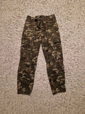 Camouflage cargopants från Amisu - Säljer ett par snygga cargopants från Amisu i storlek 34. Byxorna har klassiskt camouflage-mönster i gröna och bruna toner, flera praktiska fickor på benen och knytning i midjan. Perfekt för dig som gillar streetstyle och utility-vibe.