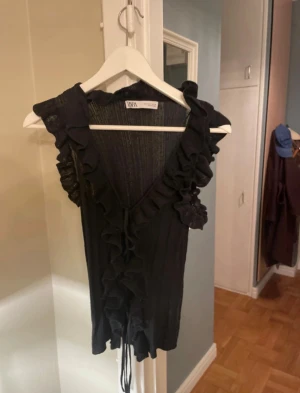 Svart volangtop från Zara - Supersnygg svart topp från Zara med volangdetaljer längs v-ringningen och framtill. Toppen är ärmlös, ribbad och har knytband framtill. Perfekt för dig som gillar en feminin och trendig look. 