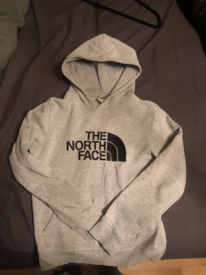 Grå hoodie från The North Face  - Säljer en grå hoodie från The North Face i storlek xxs och passar barn 10-12 åldern . Tröjan har klassisk huva, stor svart logga på bröstet och en skön, mjuk insida. Perfekt för chill dagar och streetwear-stil. Fickor framtill och ribbade muddar vid ärmslut och nederkant.