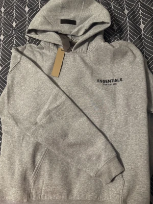 Grå Essentials Fear of God hoodie - Säljer en hoodie från Essentials Fear of God i färgen Light Oatmeal med svart logotyptryck både fram och bak.Helt ny hoodie med alla lappar kvar. Tröjan har huva, ribbade muddar och en klassisk loose/ oversized passform. Materialet är mjuk bomull och polyester, perfekt för chill dagar. 