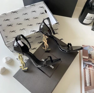 Svarta Saint Laurent klackar med YSL-detalj - Säljer ett par svarta klackar från Saint Laurent med ikonisk YSL-logga i guld som klack. Skorna har smal rem över tårna, ankelrem med spänne och är tillverkade i glansigt skinn. Perfekta för dig som vill sticka ut med lyxig och edgy vibe.