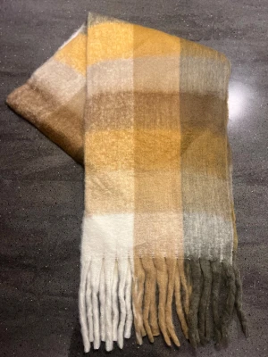 Halsduk - Supermjuk och fluffig halsduk som liknar acne studios med rutigt mönster i vitt, beige, grått, brunt och gult. Långa fransar i ändarna ger en chill vibe. Perfekt att svepa runt halsen för extra värme under kyliga dagar. Materialet känns som ull eller syntet och färgerna är harmoniska och jordnära.