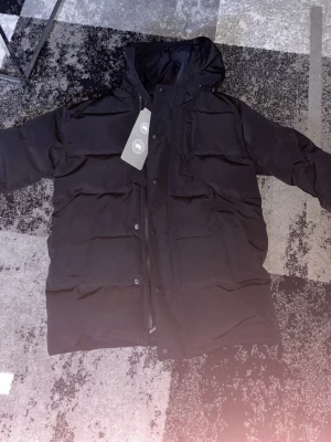 Svart pufferjacka från Canada Goose - Svart pufferjacka från Canada Goose med huva och klassisk logotyp på ärmen. Jackan har dragkedja och knappar framtill, flera fickor och är fodrad med dun. Perfekt för kalla dagar och har en clean, stilren look.