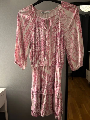 Rosa och vit mönstrad klänning från Zara - Säljer en söt kortklänning från Zara i rosa och vitt med blommigt mönster. Klänningen har trekvartsärmar, volangdetaljer och knytning vid halsen. Den är tillverkad i ett mjukt bomullsmaterial och har en avslappnad passform med resår i midjan.