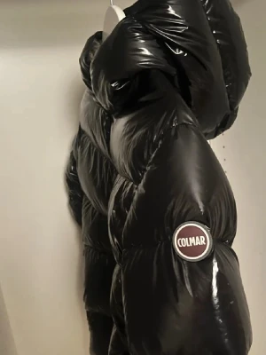 Svart Colmar pufferjacka med huva - Säljer en svart pufferjacka från Colmar med glansig finish och stor, mysig huva. Jackan har en tydlig Colmar-logga på ärmen och dragkedja framtill. Perfekt för kalla dagar och har en riktigt cool, puffig look som sticker ut. Nypris ca 5000