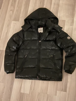 Moncler Maya - Säljer en svart pufferjacka från Moncler med huva och coolt Moncler-märke på ärmen. Jackan har dragkedjor på fickorna och quiltad design som ger en schysst look. Insidan har en unik serietidningsetikett. Perfekt för dig som vill ha en varm och snygg jacka.