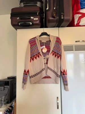 Odd Molly beige kofta med mönster - Snygg beige kofta från Odd Molly med färgstarkt mönster i lila, rött och brunt. Koftan har långärmad design, stickad struktur och knyt framtill. Detaljerad med ribbad kant och coola grafiska mönster på ärmar och framstycke. Perfekt för dig som gillar unika plagg! Från märket Odd Molly. Storlek 3 som enligt deras storleksguide motsvarar en L. Helt ny med tags/ prislapp fortfarande kvar.