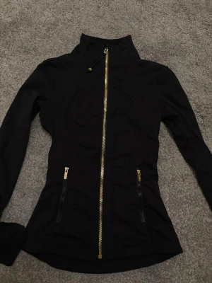 Svart define jacket från Lululemon med guld-detaljer - Snygg svart jacka från Lululemon med guld-dragkedjor och logga. Figurnära passform, hög krage och tumhål vid ärmslut. Perfekt för träning eller chill, tillverkad i stretchigt syntetmaterial som andas. Flera praktiska fickor med dragkedja. I storlek 2