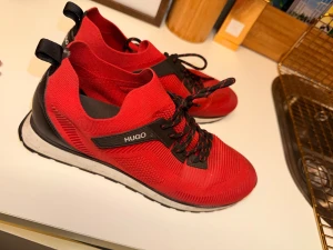 Röda sneakers från Hugo Boss - Snygga röda sneakers från Hugo Boss med svart och vit sula, svarta detaljer och snörning. Ovandelen är i textil med sportig känsla och logga på sidan samt under sulan. Perfekta för dig som vill sticka ut med stilrena och moderna skor.