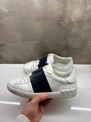 Valentino Garavani Open sneakers vit/marin - Snygga Valentino Garavani Open sneakers i vitt skinn med bred marinblå läderdetalj över mitten. Klassisk rund tå, vita snören och ikoniska nitar baktill på sulan. Perfekt för dig som vill ha en clean men lyxig look.