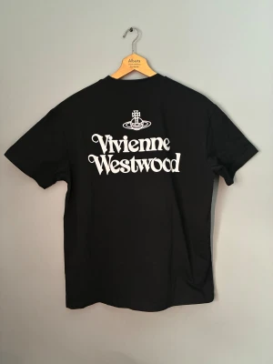 Svart t-shirt Vivienne Westwood - Svart t-shirt från Vivienne Westwood med stor vit logotyp på ryggen och mindre logga framtill. Klassisk rund hals och korta ärmar. Tillverkad i ekologisk bomull, perfekt för dig som gillar ikoniska streetwear-plagg med tydlig branding.