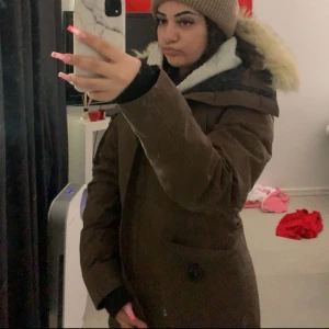 Brun parkas från Canada Goose med päls - Säljer en brun parkas från Canada Goose med fluffig päls på huvan, patch på ärmen och flera fickor med både dragkedja och knappar. Jackan har lång ärm med ribbad mudd och är perfekt för kalla vinterdagar. Snygg och funktionell design med klassisk look.