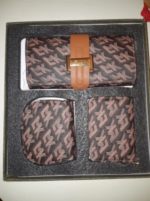 Set med plånbok och accessoarer - Snyggt set med plånbok, kortfodral och myntbörs från EMN. Alla delar har ett brunt och svart geometriskt mönster med rosa detaljer och en guldfärgad metallspänne på plånboken. Materialet är syntet och designen är modern och stilren.