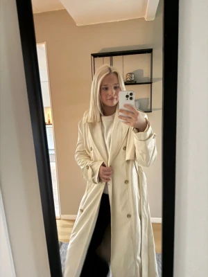 Beige trenchcoat med bälte - Lång beige trenchcoat med en skinn material , knappar framtill och bälte i midjan. Jackan har snygga detaljer vid axlarna och ärmarna.