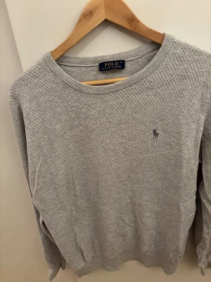 Grå stickad tröja Polo Ralph Lauren - Snygg grå stickad tröja från Polo Ralph Lauren i storlek M. Tröjan har ett diskret mönster och den klassiska lilla loggan broderad på bröstet. Rund hals och långa ärmar, perfekt för dig som gillar stilrena och tidlösa plagg.