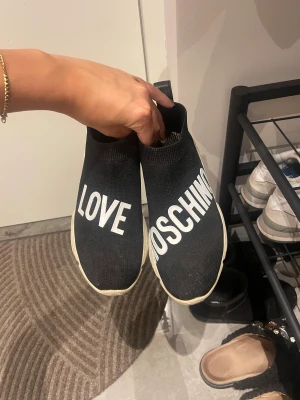 Love Moschino svarta sneakers med hjärta - Svarta sneakers från Love Moschino med vit text på ovansidan och chunky vit sula. Stickad ovandel i syntetmaterial, röda hjärtan bak på hälen och under sulan. Snygg och unik design med platt sula och rund tå. Perfekt för dig som gillar statement pieces.